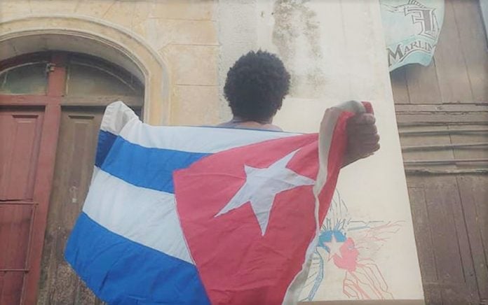 El artista Luis Manuel Otero Alcántara sostiene una bandera cubana frente a una cámara de vigilancia policial