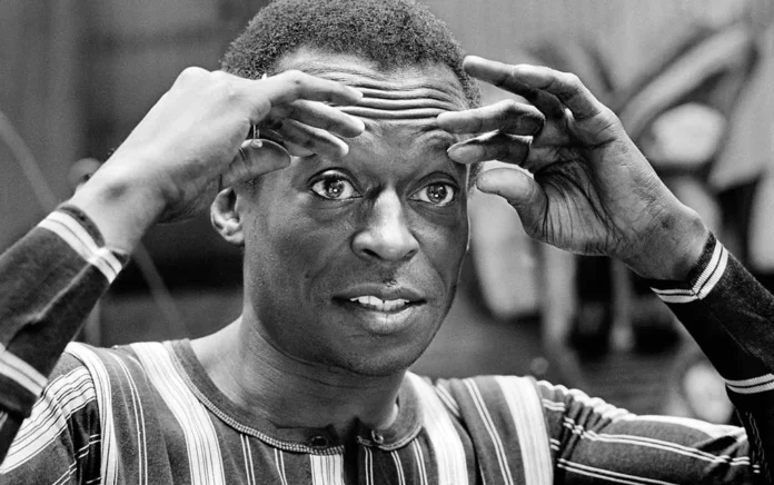 Retrato en blanco y negro del trompetista Miles Davis enmarcando su rostro con ambas manos y mirada intensa, en un estudio de grabación.