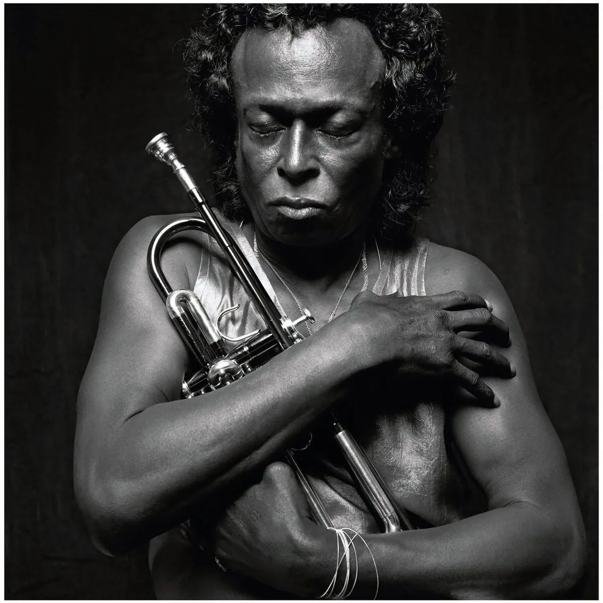 Retrato en blanco y negro de Miles Davis con el torso desnudo, los ojos cerrados, abrazando su trompeta contra el pecho sobre fondo negro.