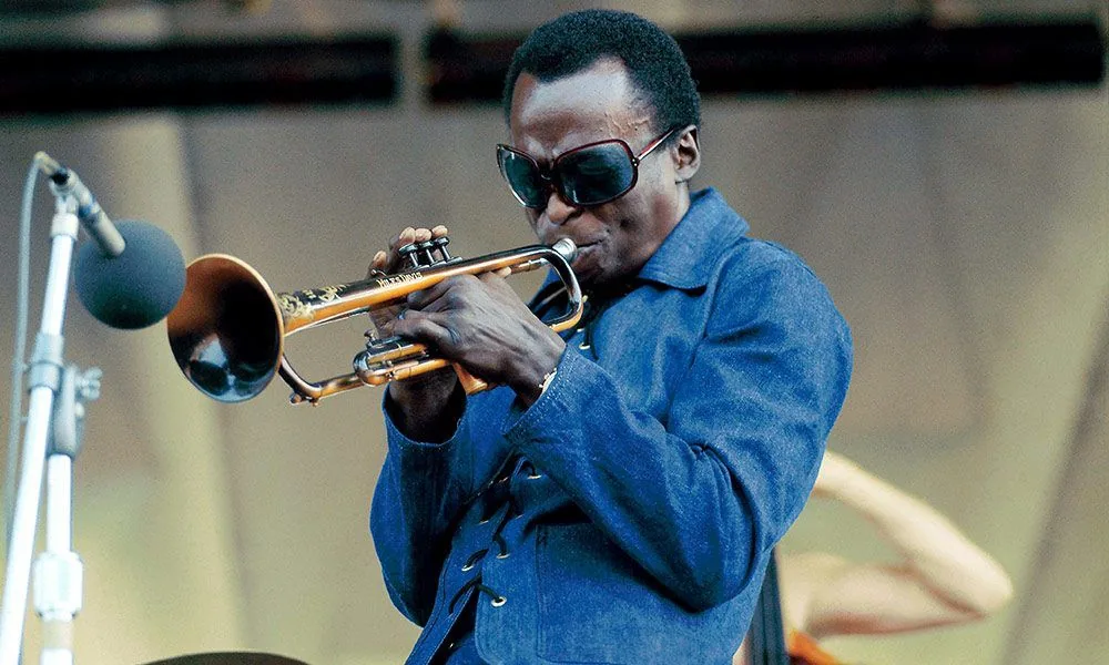 Miles Davis tocando la trompeta en vivo, con chamarra de mezclilla y lentes oscuros, durante el Newport Jazz Festival de 1969.