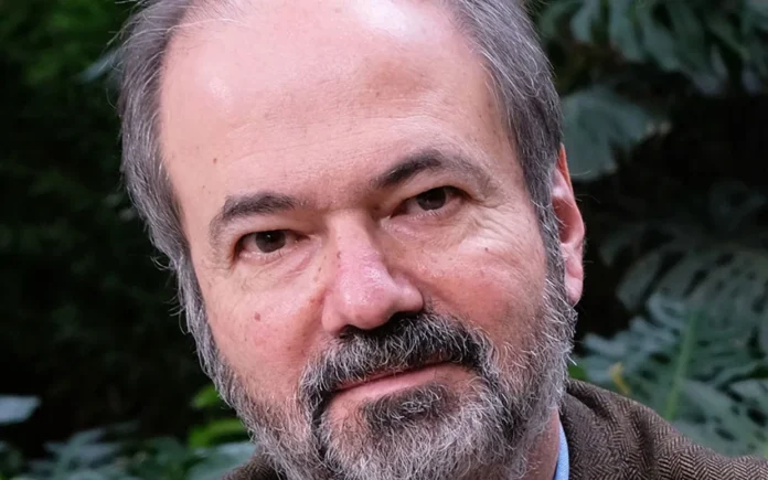 Retrato editorial de un hombre de cabello y barba grises con blazer marrón, identificado como el escritor Juan Villoro, ganador del Premio Carlos Fuentes 2026.
