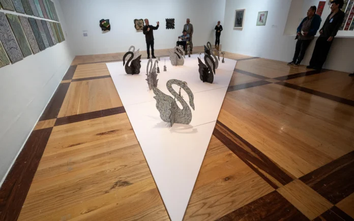 Sala de exposición con una instalación de esculturas de cisnes sobre una plataforma blanca y varias personas observando obras en las paredes.