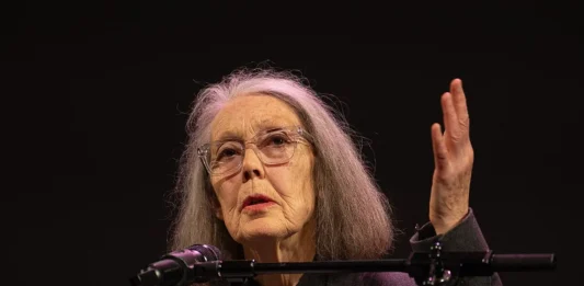 Anne Carson, de cabello gris y gafas, habla desde un atril con micrófono durante un acto cultural.