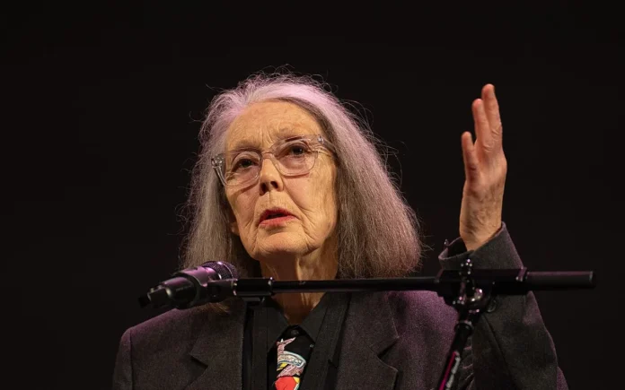 Anne Carson, de cabello gris y gafas, habla desde un atril con micrófono durante un acto cultural.