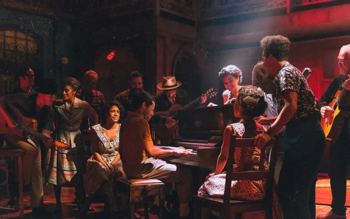 Ensamble de músicos y actores en escena alrededor de un piano durante una función del musical ‘Buena Vista Social Club’, bajo iluminación cálida rojiza.