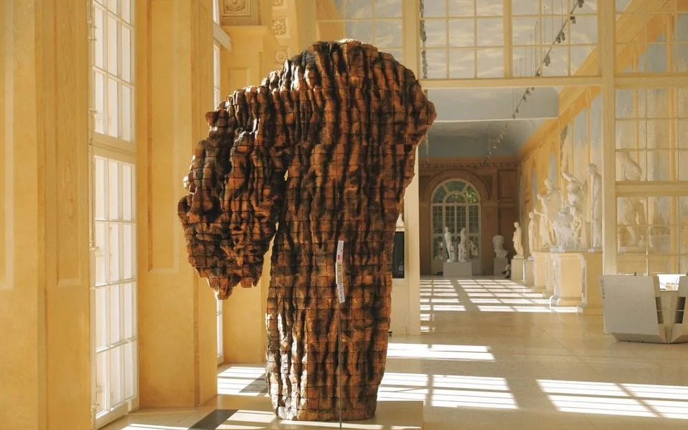 Escultura voluminosa de Ursula von Rydingsvard, de textura de tiras apiladas en tonos marrones y negros, exhibida en una sala clásica con ventanales.