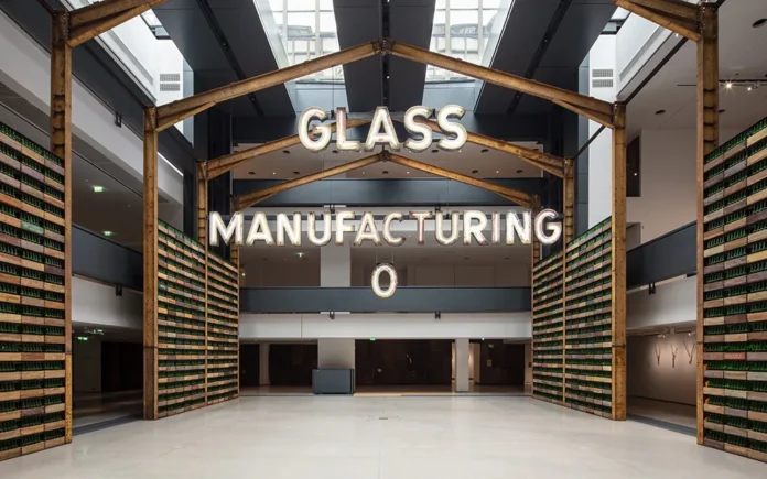 Instalación de Ibrahim Mahama en la Fondation Cartier: acceso enmarcado por vigas metálicas oxidadas, paredes hechas de botellas de vidrio y el letrero luminoso “Glass Manufacturing”.