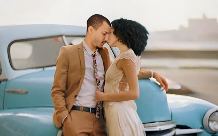 Pareja frente a frente apoyada en un automóvil clásico turquesa en una zona costera de La Habana, imagen promocional de ‘Havana Coyotes’.